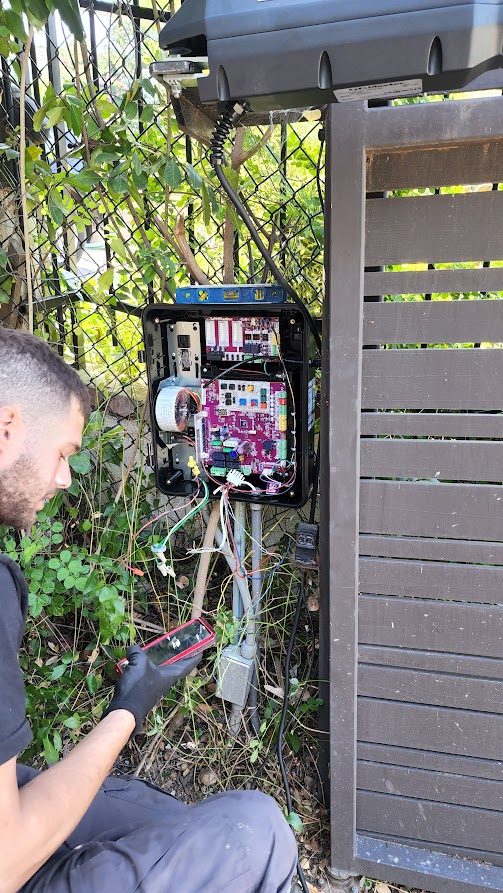 Technician kneeling at an open gate operator cabinet, testing the control board with a multimeter