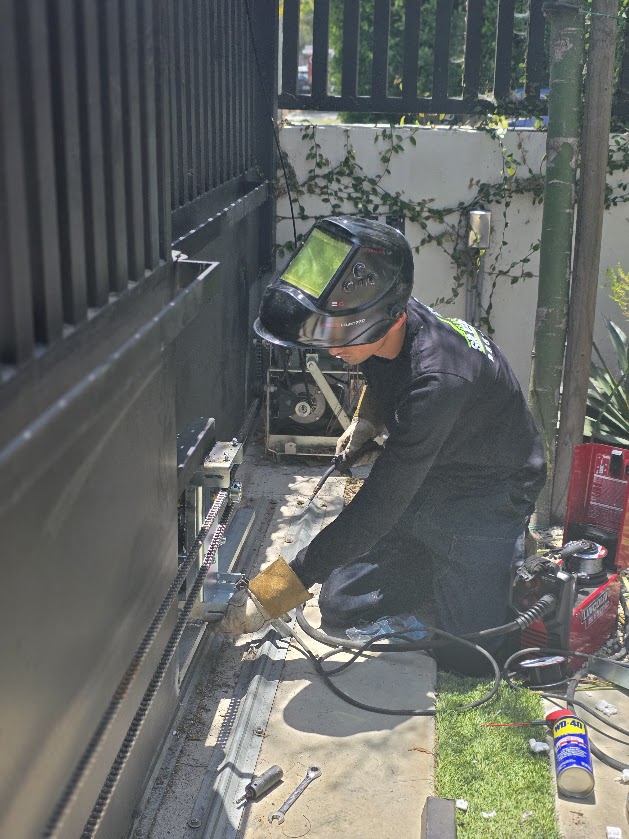 Technician in a welding helmet kneeling at the base of a dark slatted gate, welding the track back into place