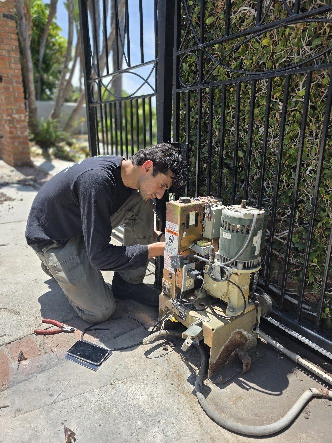 Technician working on an exposed gate operator at a wrought iron gate, with the motor, gearbox and chain drive open in front of him