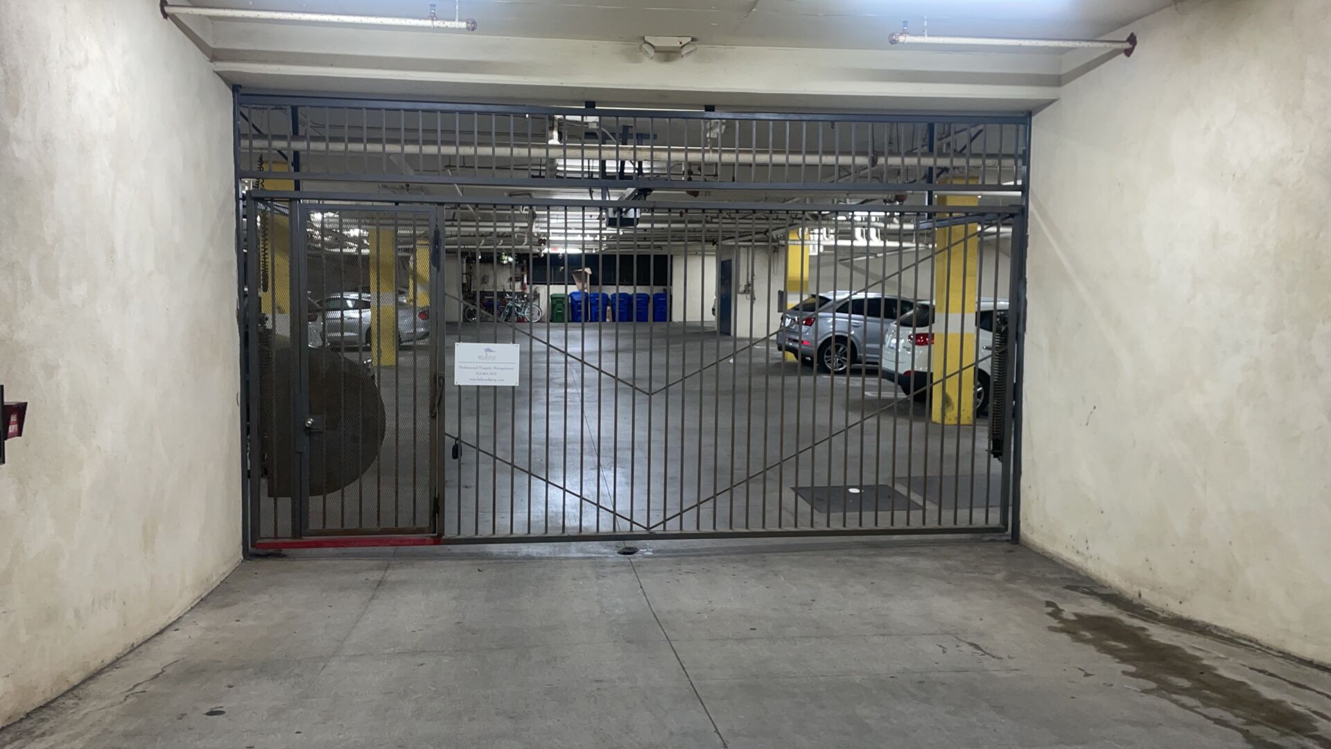 Barred steel security gate across the entrance to a subterranean parking garage, with parked cars and yellow columns visible inside