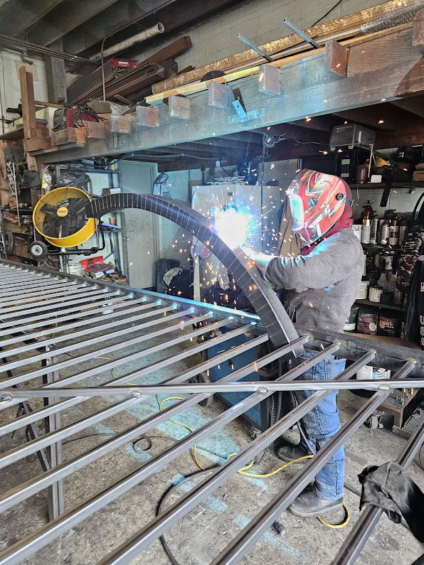 Fabricator in a welding helmet welding a curved steel gate frame on a jig inside the workshop, with sparks and arc flash visible